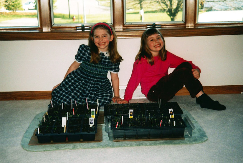 Two kids sit behind a box of seedlings in front of a window.