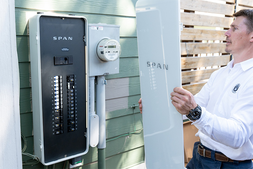 A man holds the cover of a power panel next to the panel hanging on the outside of a home.