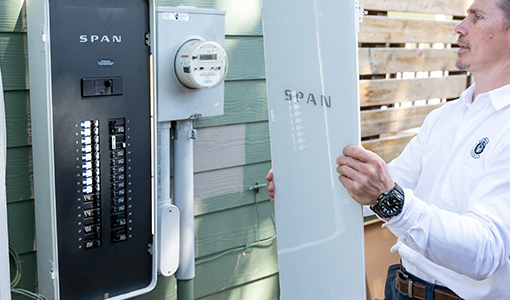 Continue reading about A man holds the cover of a power panel next to the panel hanging on the outside of a home.
