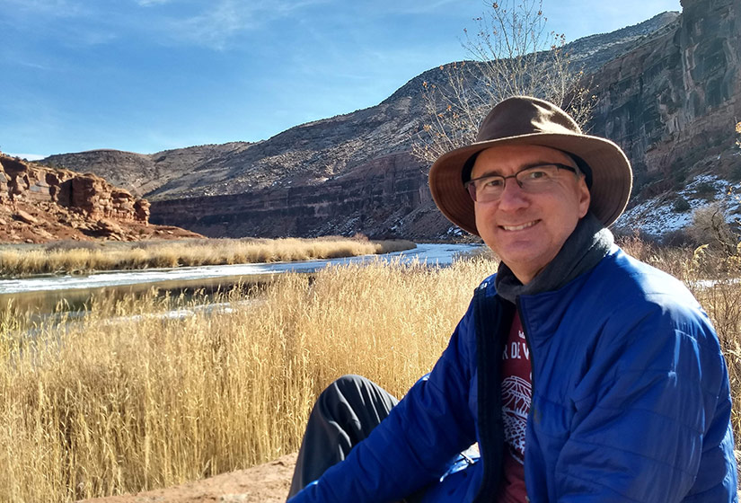 A man sits on a river bank with cliffs in the background.