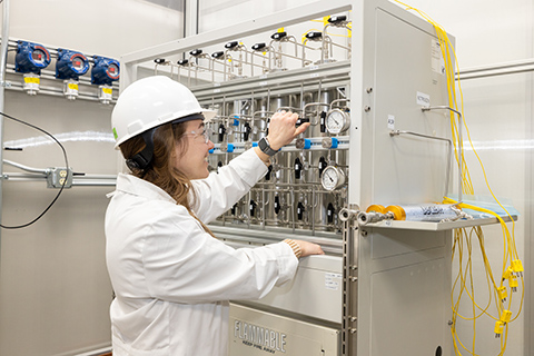 A person in a lab coat and hard hat places their hands on a machine in a lab.