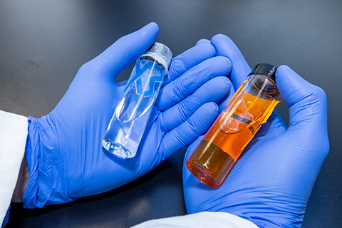 Two gloved hands hold a pair of vials, one with clear liquid and one with amber liquid.