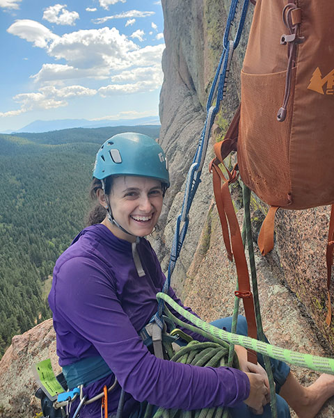 A person wearing rock climbing equipment and suspended from a rock using ropes smiles.