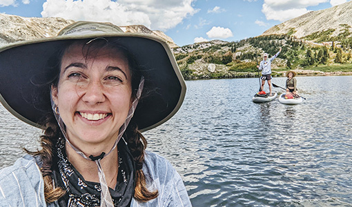 Continue reading about A person wearing a hat taking a selfie in front of two paddleboarders on a body of water.