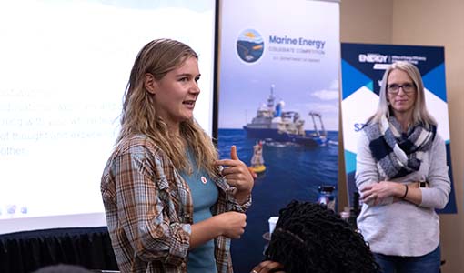 Continue reading about A person speaks in front of posters and a presentation screen as another person watches.