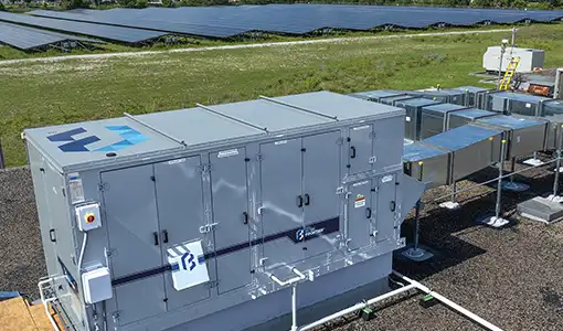 Continue reading about Photo of a large metal air conditioning unit with vents and access panels. The unit is adjacent to a field of photovoltaic panels.