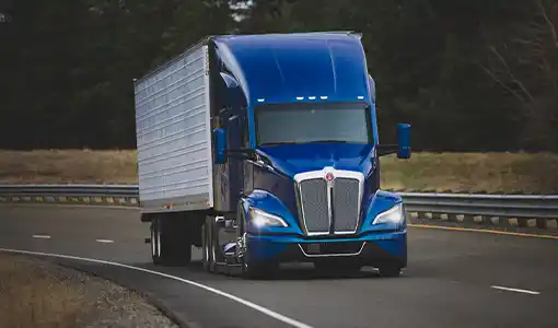 Continue reading about A blue freight truck in the foreground on a highway flanked by grass and dark trees in the background.