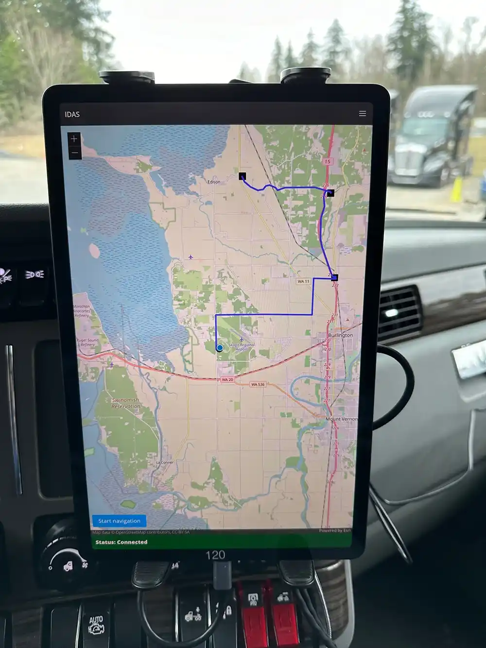 A close-up of a touchscreen tablet mounted on a vehicle’s dashboard. The tablet shows a route on a map.