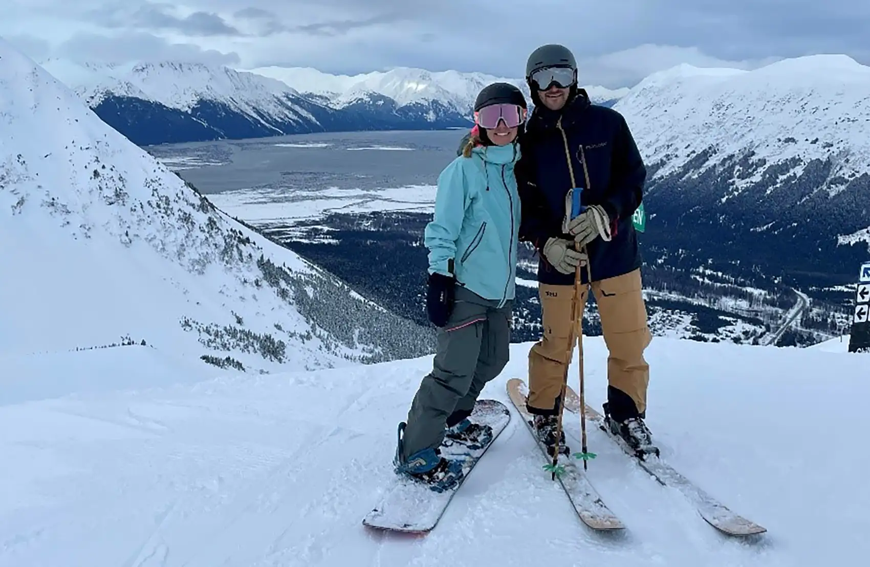 Two people in ski gear stand on a snowy slope with snow-capped mountains in the background