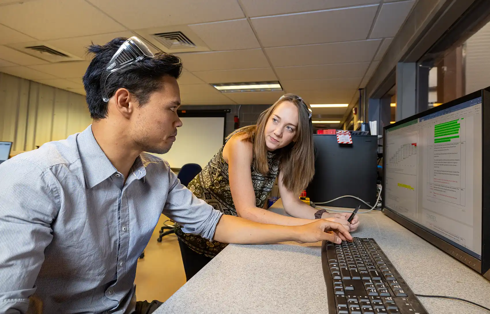 One person sits and another stands in front of a table with a computer screen displaying a graph and text.