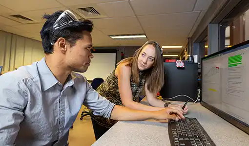 Continue reading about One person sits and another stands in front of a table with a computer screen displaying a graph and text.