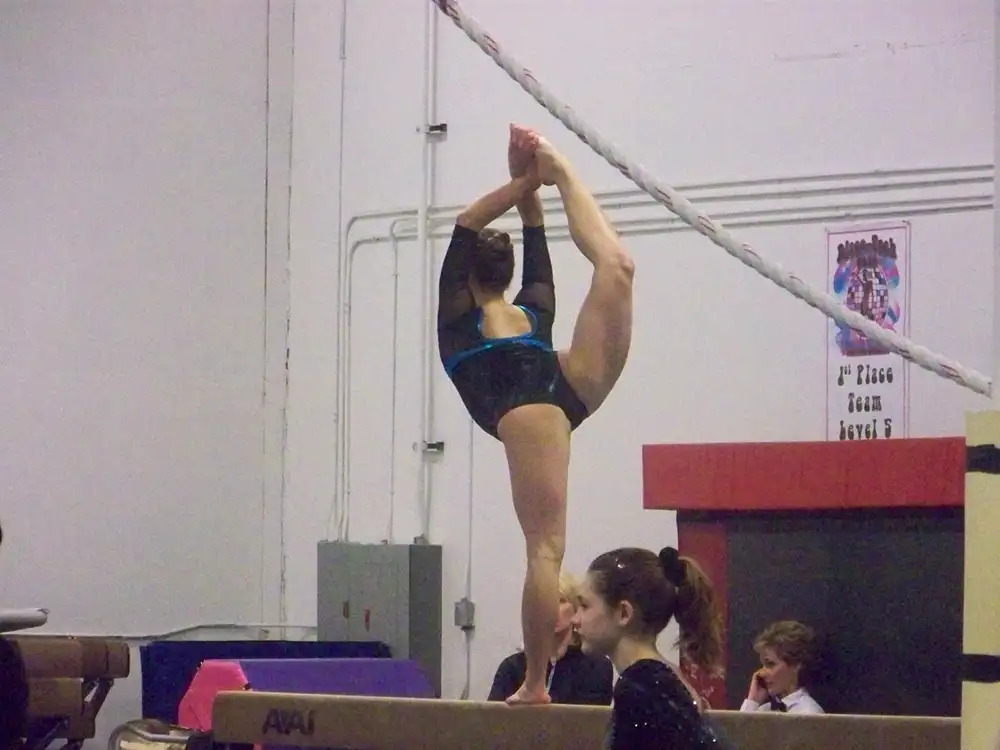 A young gymnast balancing on a balance beam while pulling one leg up behind her head