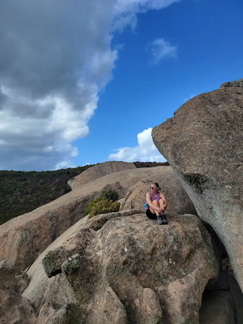 Taylor wearing sunglasses and hiking boots while sitting on top of a large rock formation