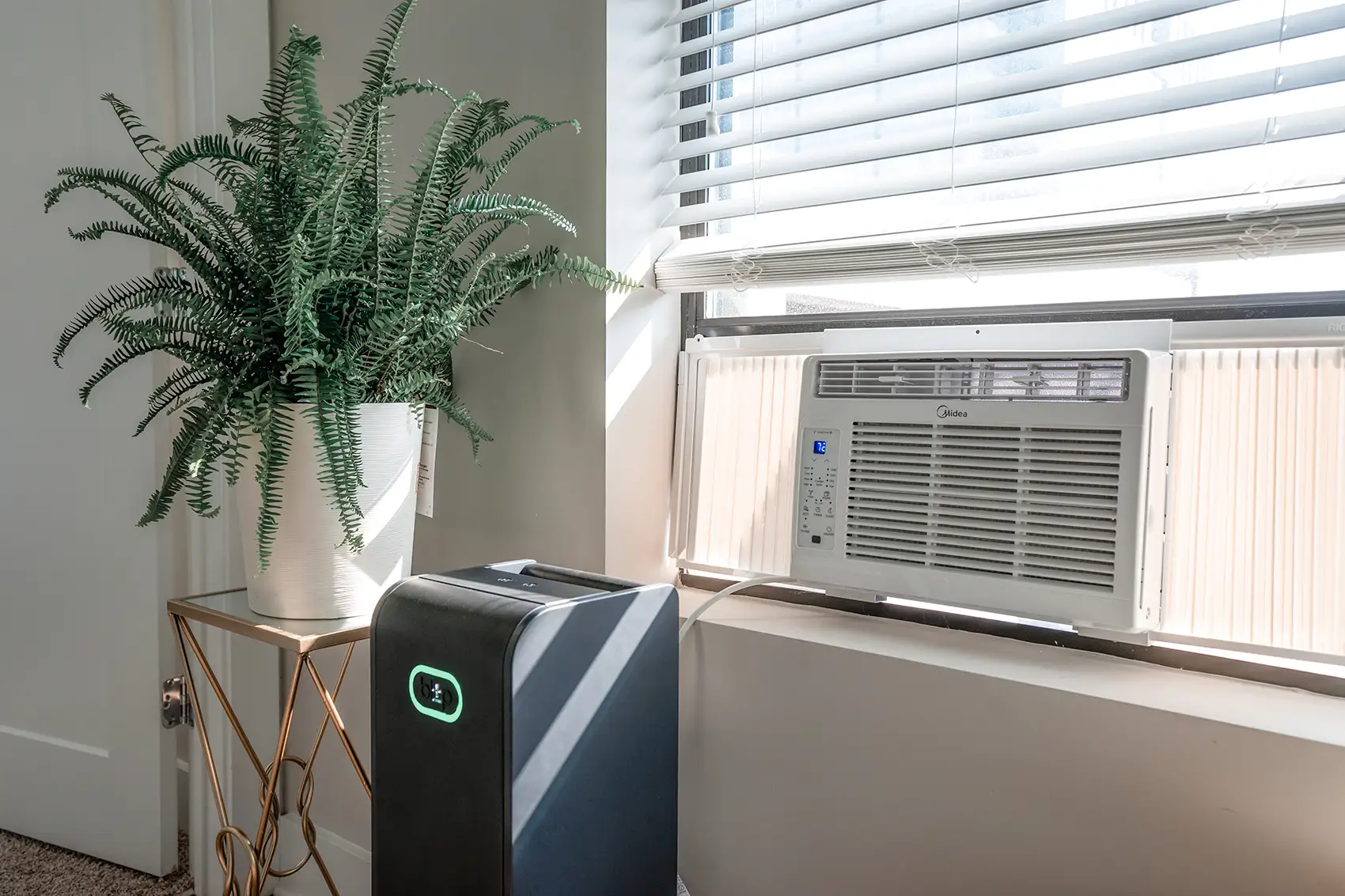 20260113-nrel-blip-energy-collaborate-on-smart-home-battery-system-1 A black smart battery sits on the floor plugged into a window air conditioner beside a window and doorframe, with a plant on a nearby table against white walls and blinds.