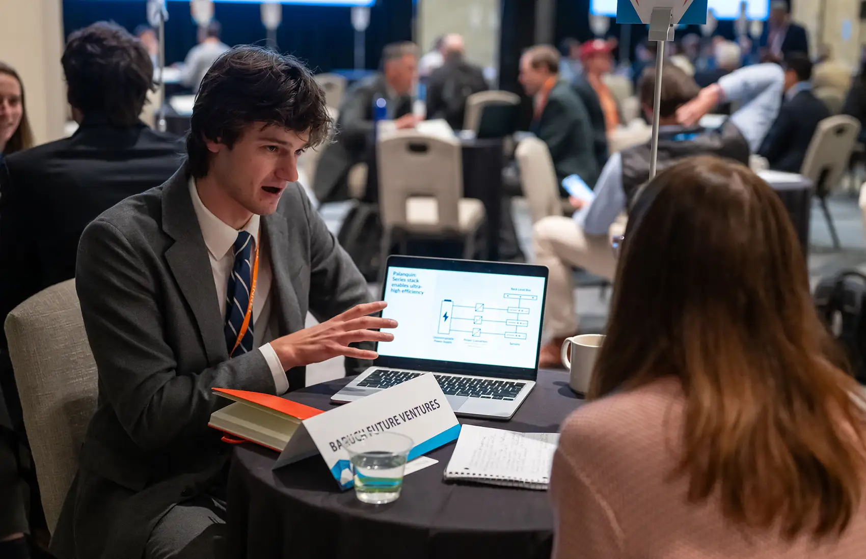 Two people sit at a table, one talking and gesturing toward a laptop screen.