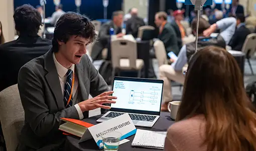 Continue reading about Two people sit at a table, one talking and gesturing toward a laptop screen.