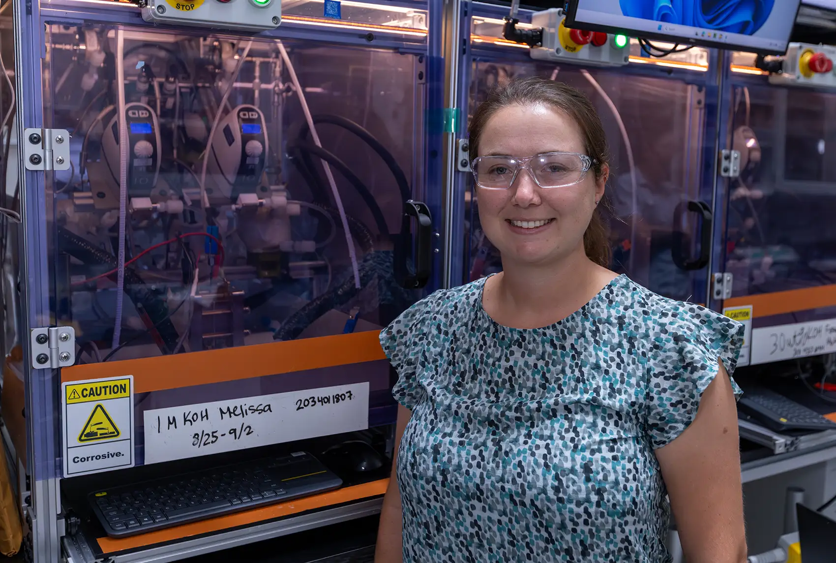 A woman with safety goggles stands in front of electrolyzer testing equipment.
