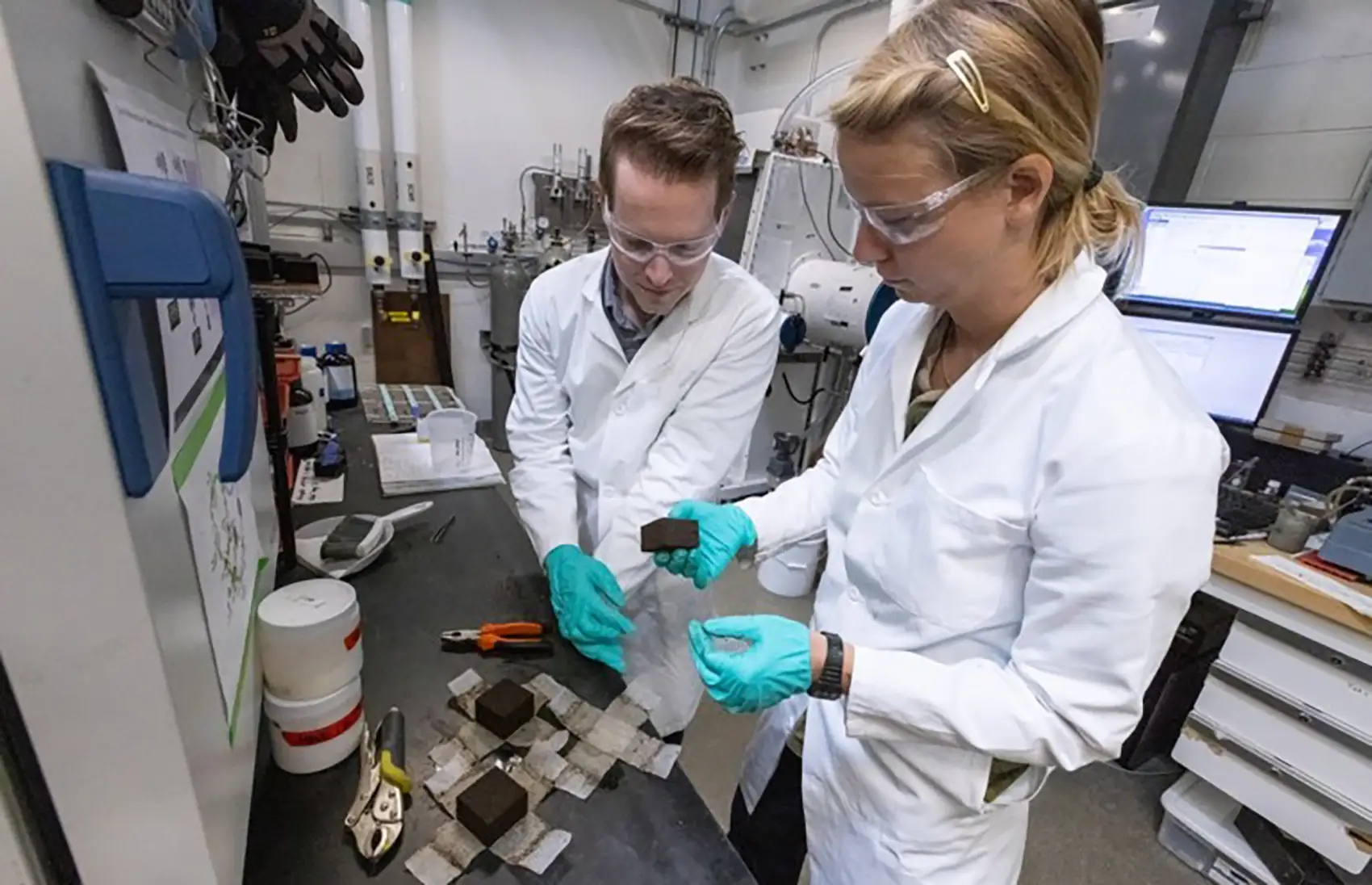 Paul wearing a lab coat, gloves, and goggles and standing next to their colleague, who picks up a material in the shape of a small brick.