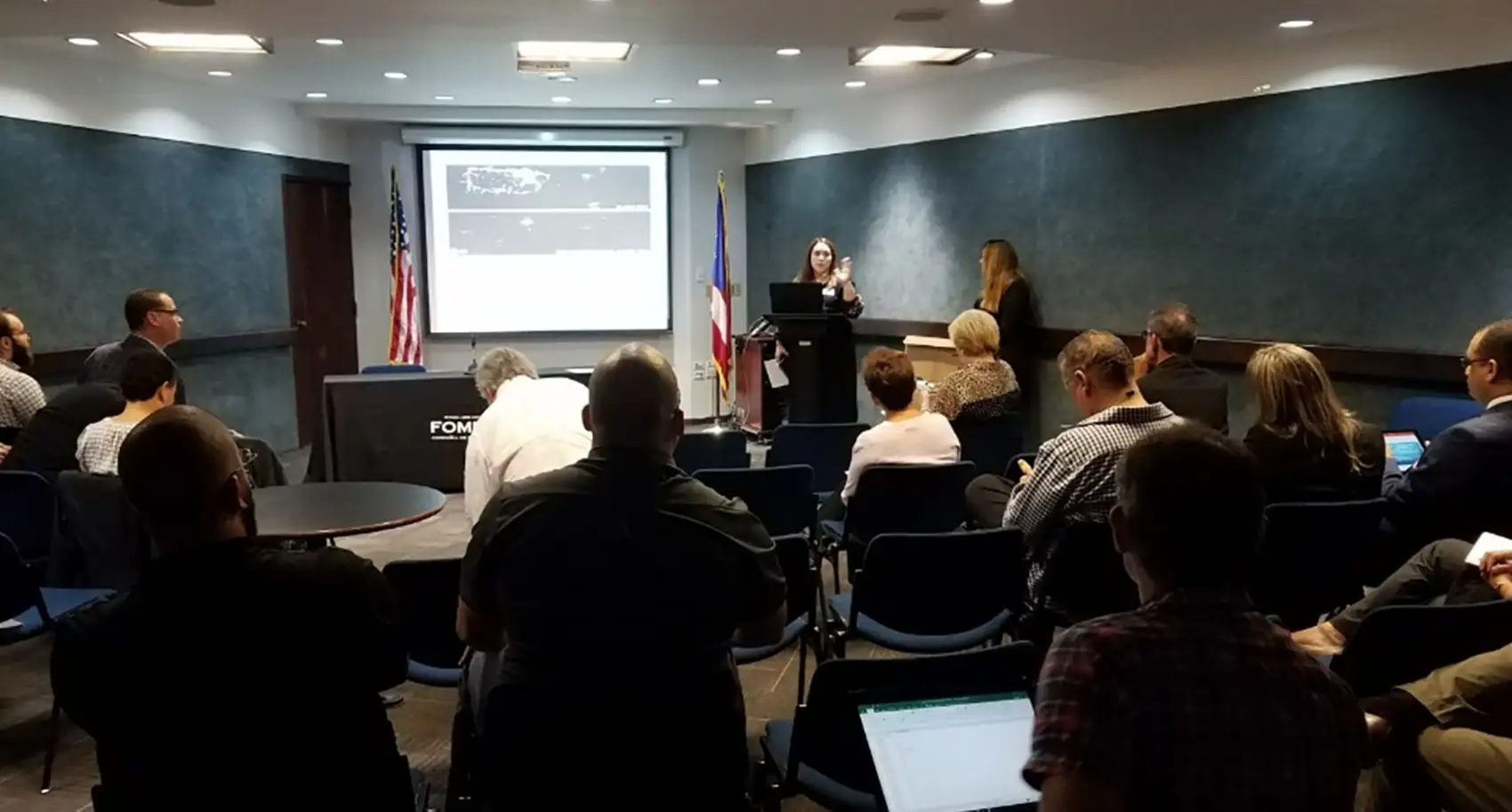 A presenter speaking to a room full of people in front of a screen.