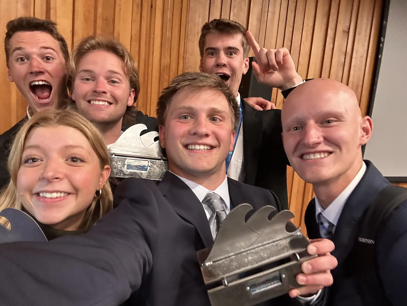 A team of six smiling student competitors pose for a selfie. Three students are holding metal trophies featuring a wave design
