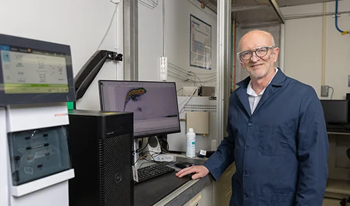 Continue reading about A man wearing a dark blue lab coat stands at a desk with a computer and machine with another screen.