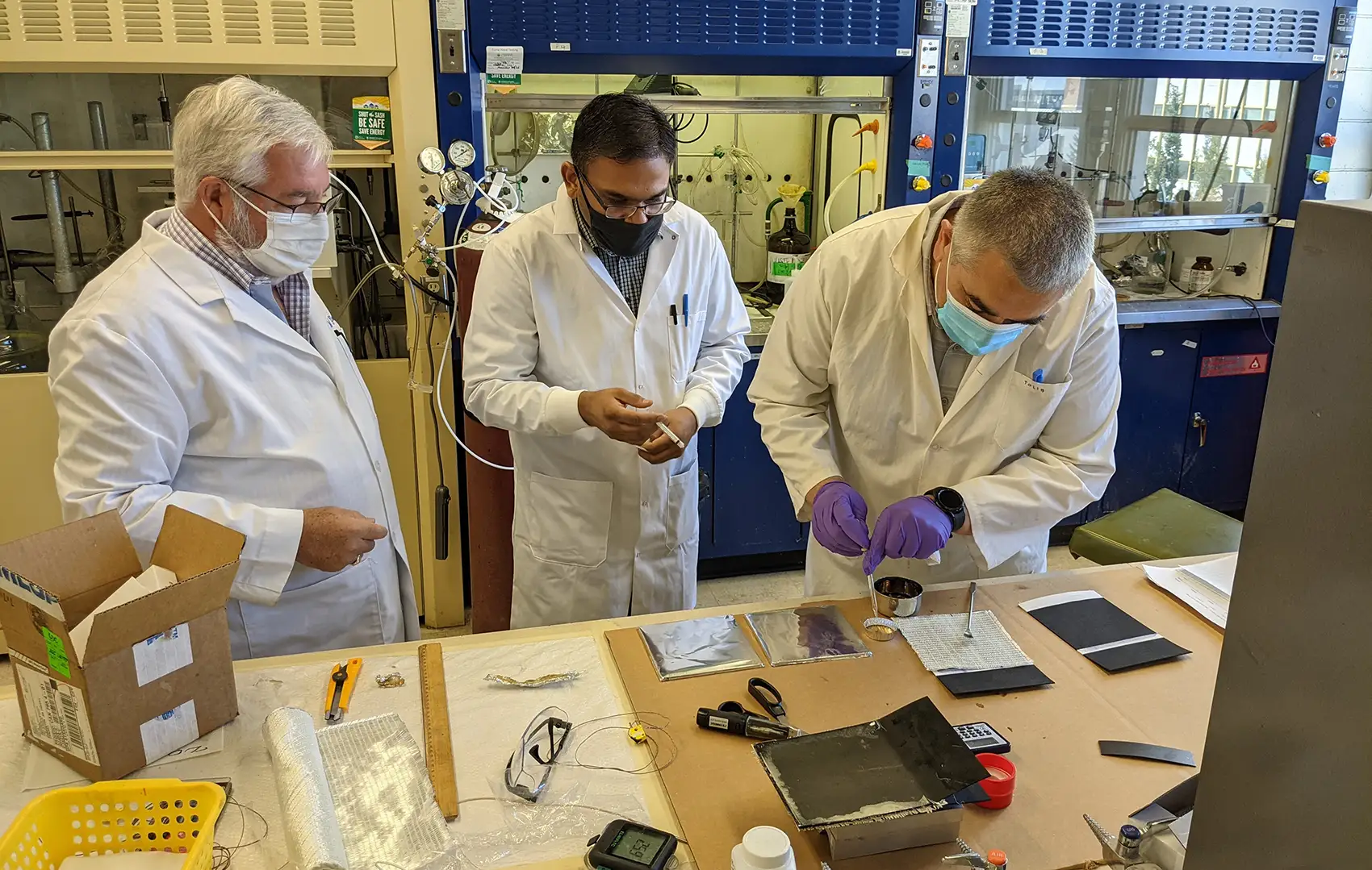 : Two people in lab coats and masks watch as a third pipes something into a container placed on a table.
