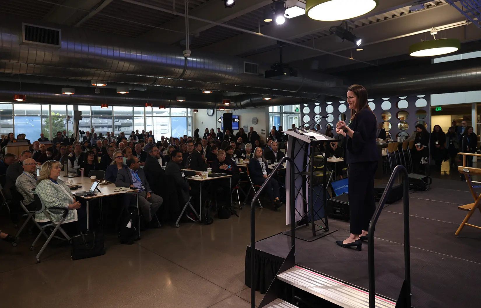 A woman stands on stage behind a podium while speaking and looking out over a large crowd gathered in a building.