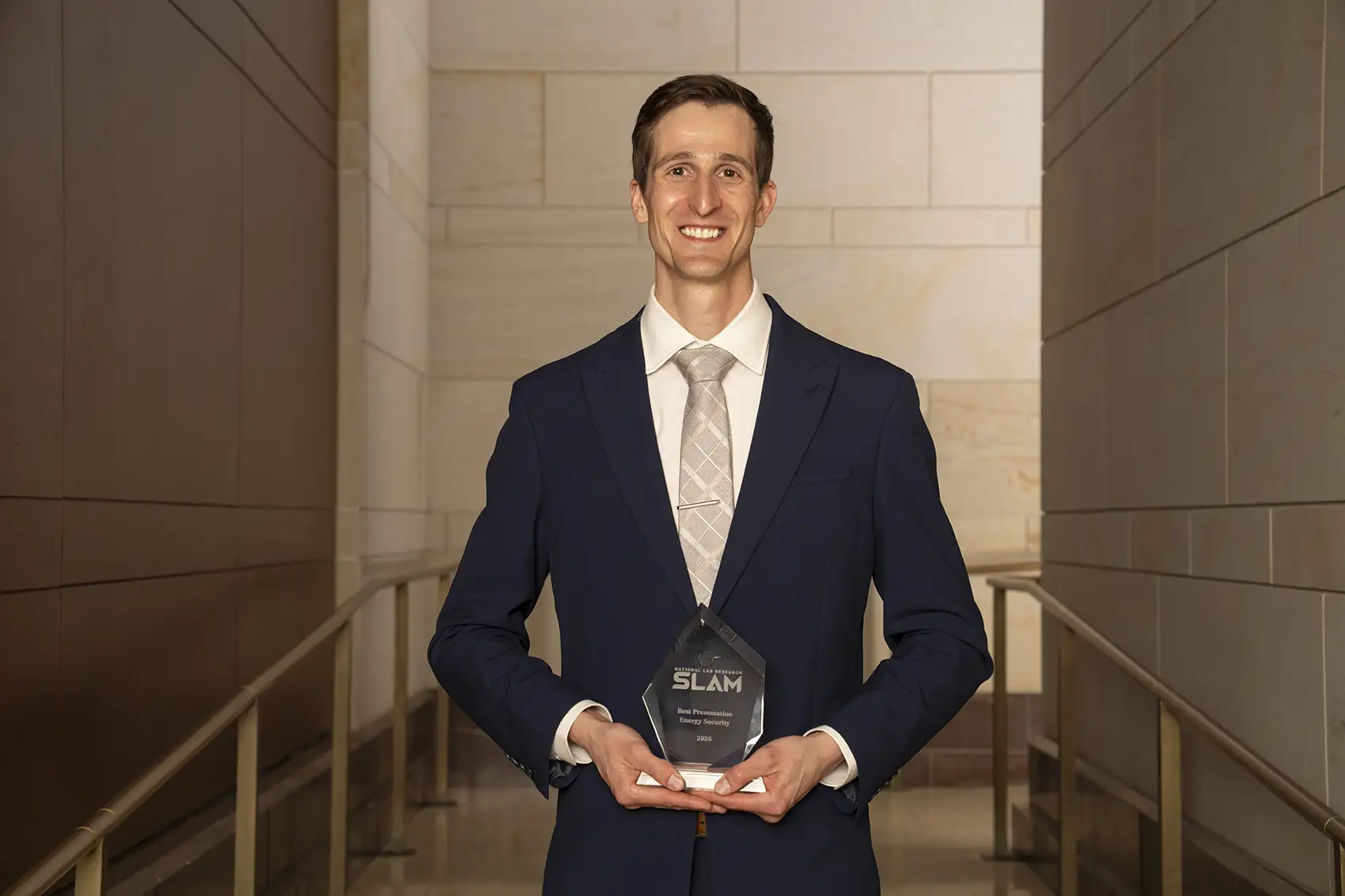 20260526-nlr-kalen-rasmussen-wins-energy-security-award-at-national-lab-research-slam-0217 A person in a suit and tie holding an award and smiling at the camera