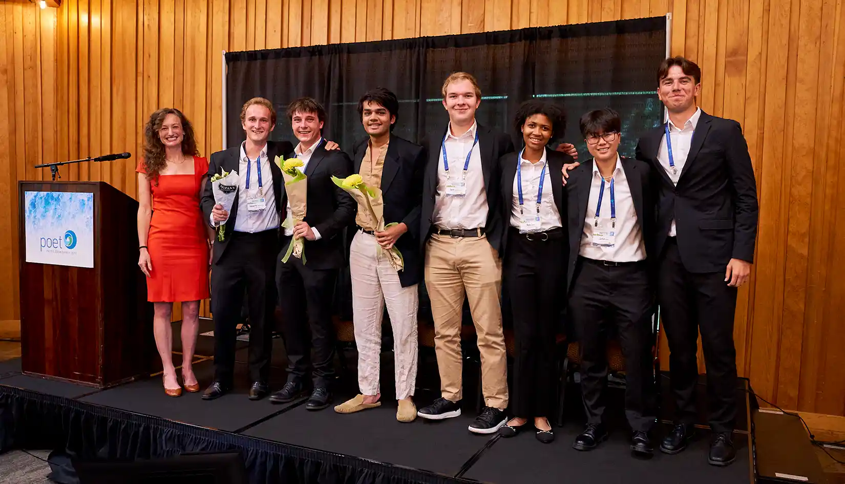 202607-mecc-winners-005 Photo of eight people, three of whom are holding bouquets, standing on a stage next to a lectern bearing the Pacific Ocean Energy Trust logo.