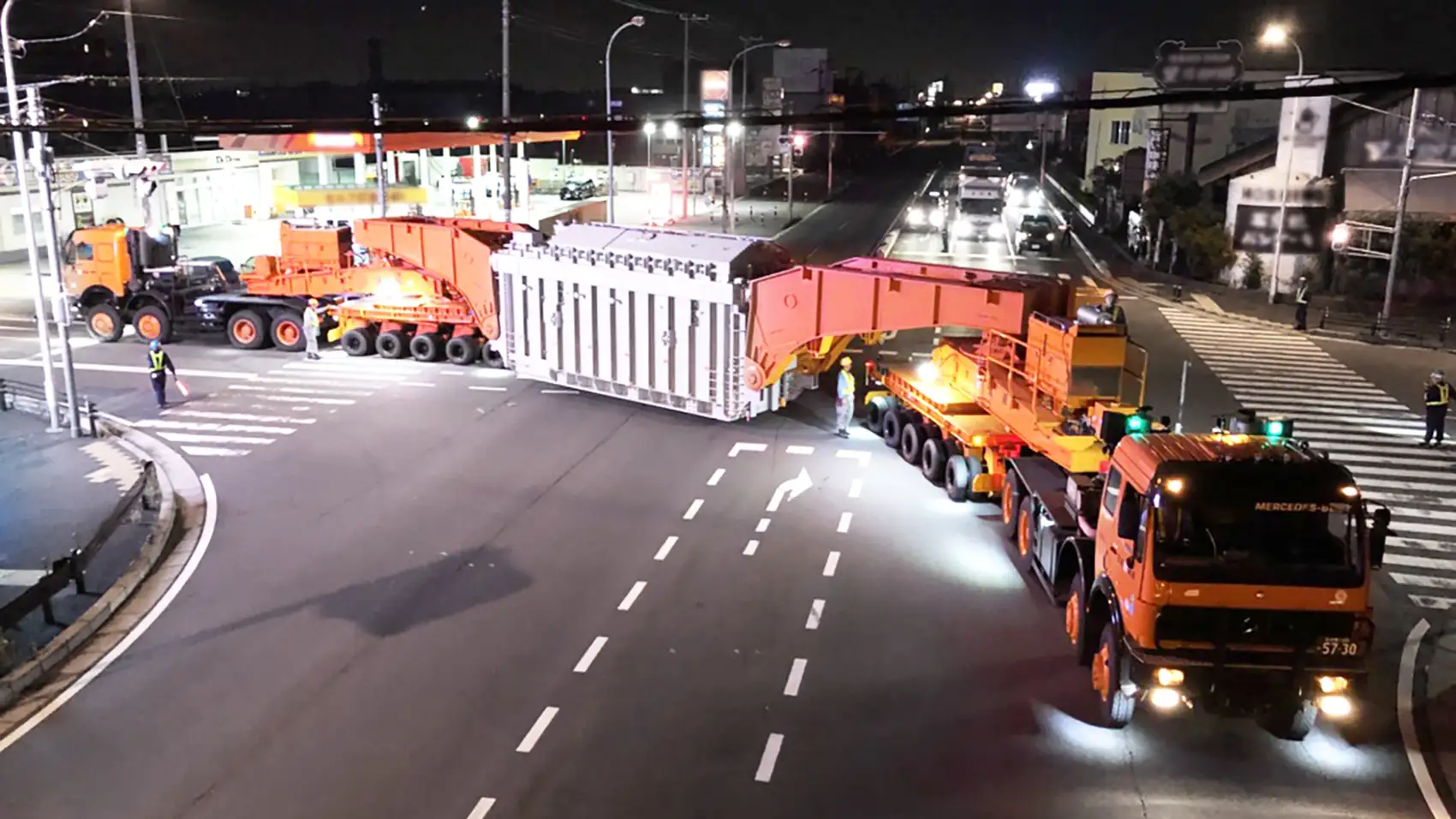 20260715-us-hydropower-faces-supply-chain-challenges-with-large-power-transformers-nippon A nighttime photo of a massive heavy-transport configuration navigating an intersection. A large, rectangular white industrial load is suspended between two oversized, multi-axle orange transport rigs.