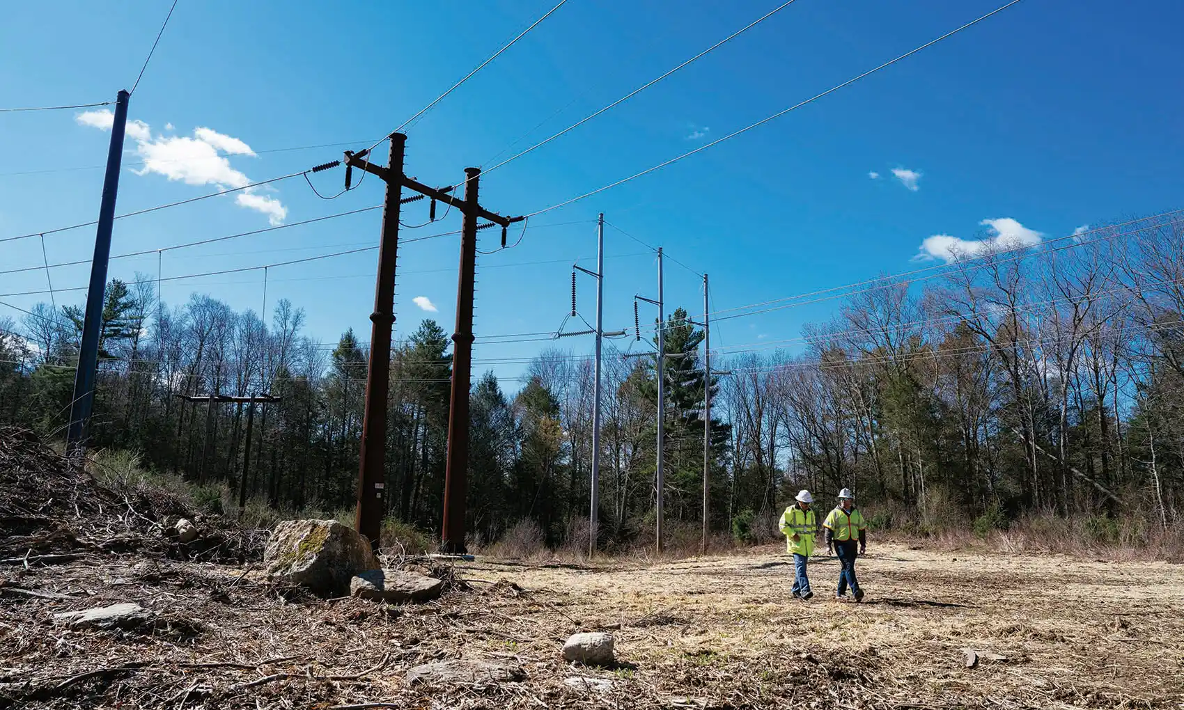 20260720-affordable-homes-cooling-centers-faster-grid-yes-please-west-charlton-substation Two utility workers walk beneath high-voltage power lines in a cleared area on a sunny day.
