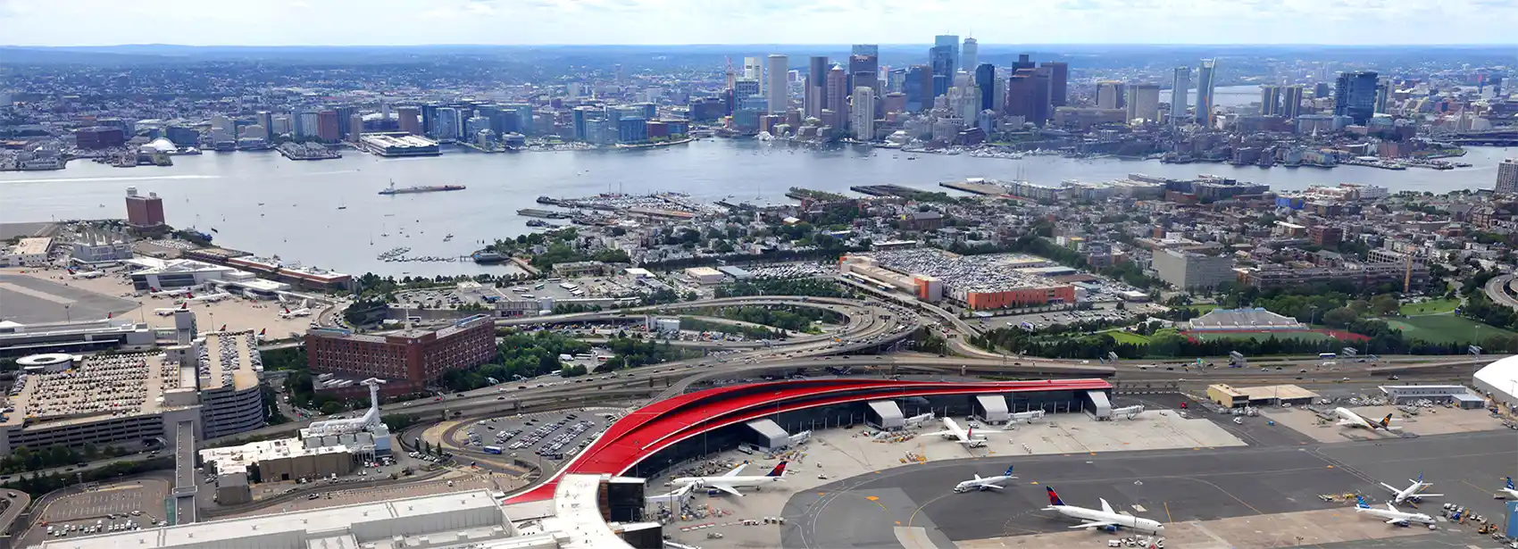 20260723-as-jet-fuel-supplies-tighten-can-other-fuels-meet-demand-massport Aerial view of an airport next to a body of water and a city skyline.