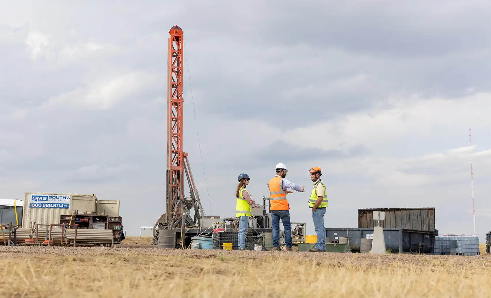 20260727-the-fast-lane-3-ways-geothermal-companies-can-get-more-heat-102003 Three people in reflective vests and hard hats stand in a field near drilling equipment