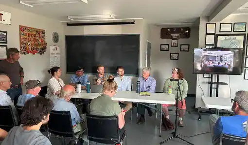 People sit in a room and face a table where six people are seated.