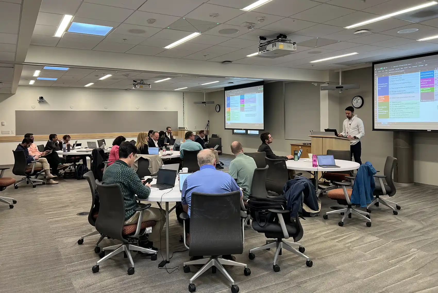 A group of people sitting around tables in a room while a person stands at a podium giving a presentation.