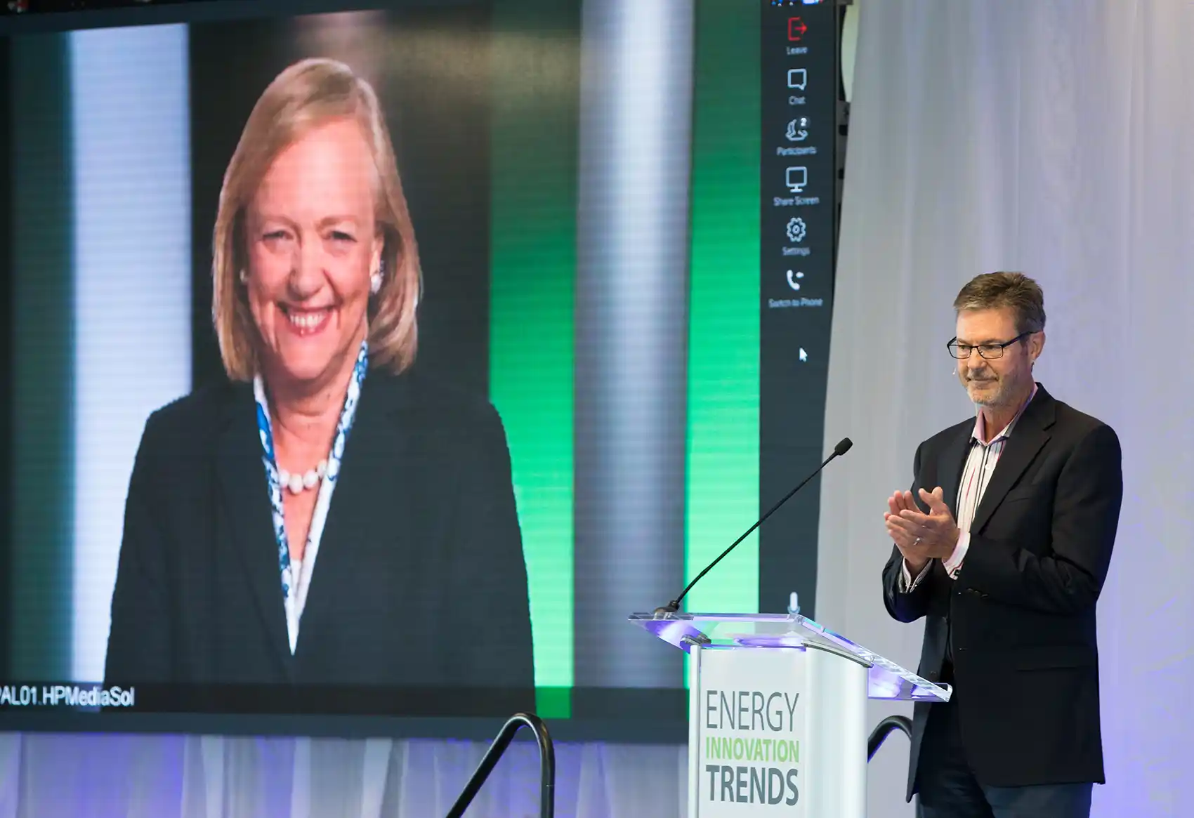 A person behind a podium clapping in front of a presentation screen with a person's face on it.