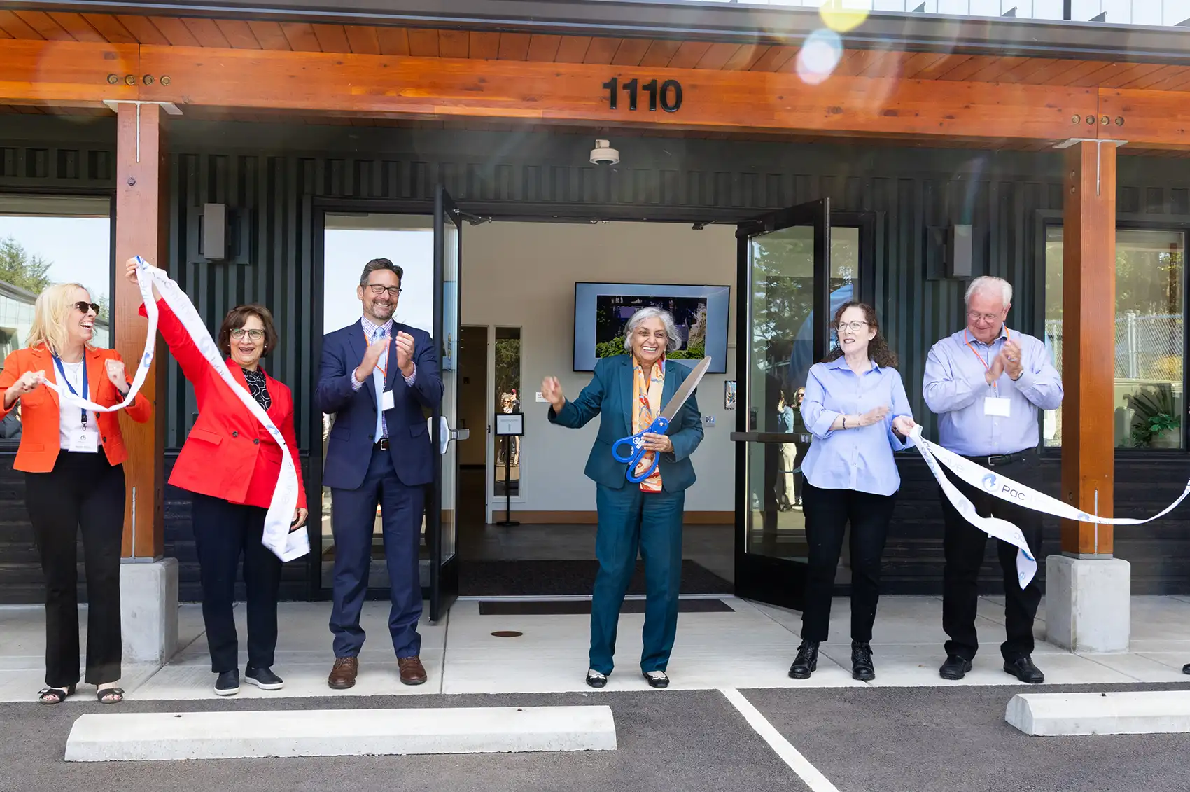 20260909-pacwave-opens-a-giant-leap-for-marine-energy-9U3A5616 A photo of six people celebrating at the front entrance of a building. In the center, a person holds oversize scissors.