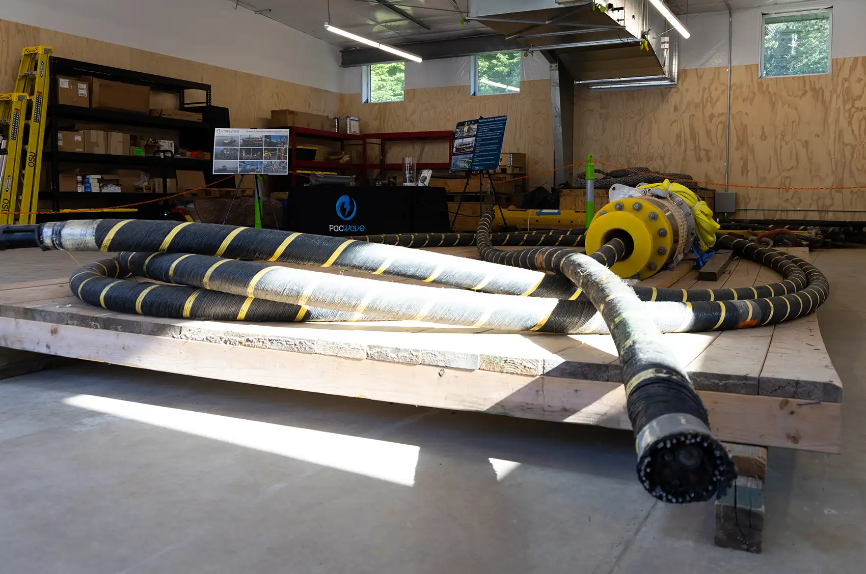 20260910-pacwave-opens-a-giant-leap-for-marine-energy-9U3A5334 A coiled section of cable sits atop of a wood pallet on the floor of a rough-framed building containing some shelving and a few signs.