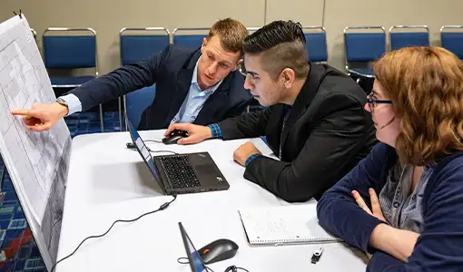 Continue reading about Three people seated at a table with laptops, a notepad, and a thumb drive study information printed on a table-length section of poster board propped up in front of their table.