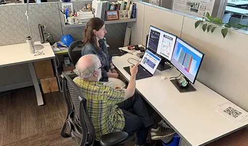 Two people sit at a desk with three computer screens.