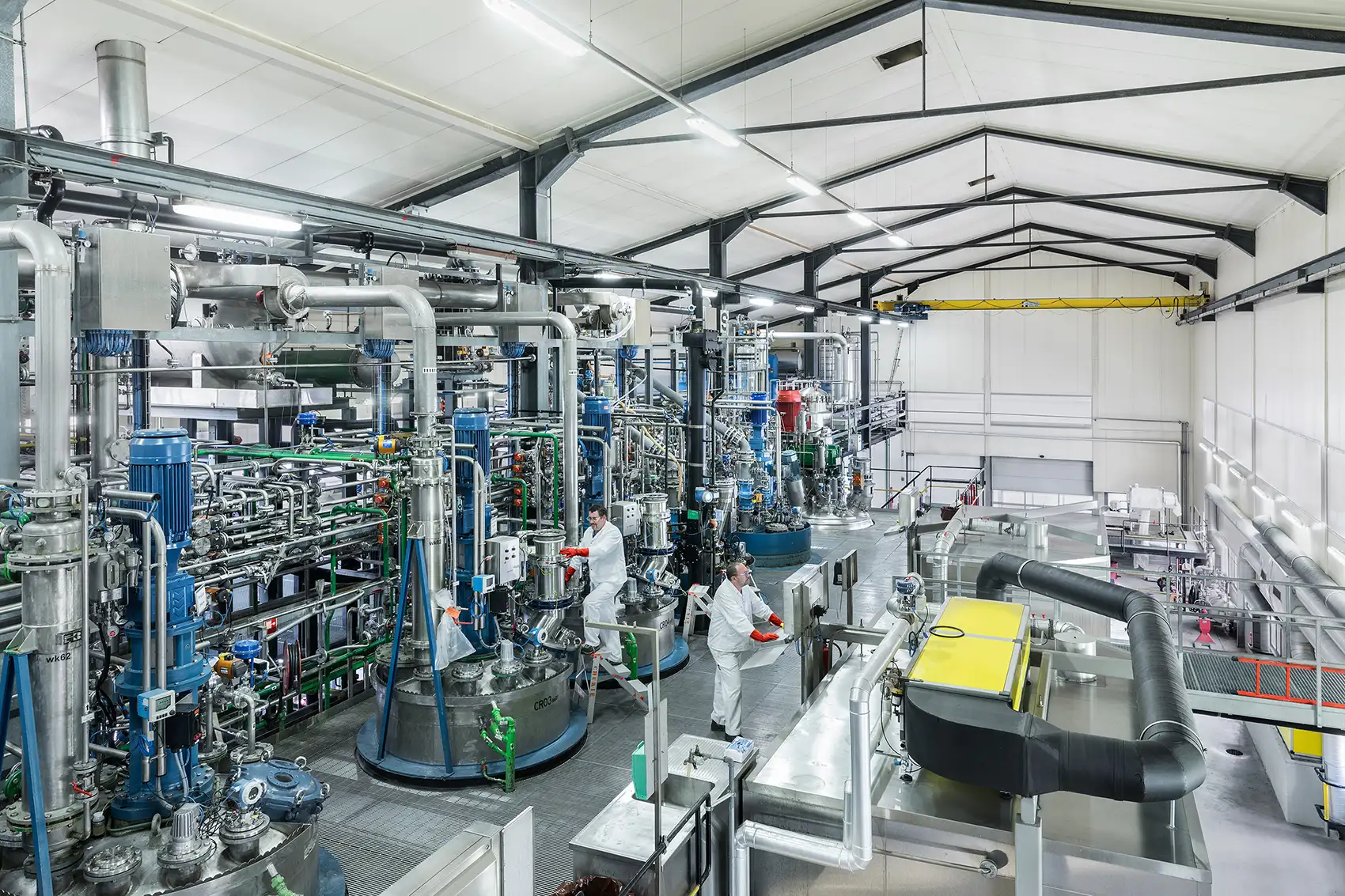 Industrial facility with stainless steel pipes, tanks, and processing equipment, with researchers in white protective clothing.