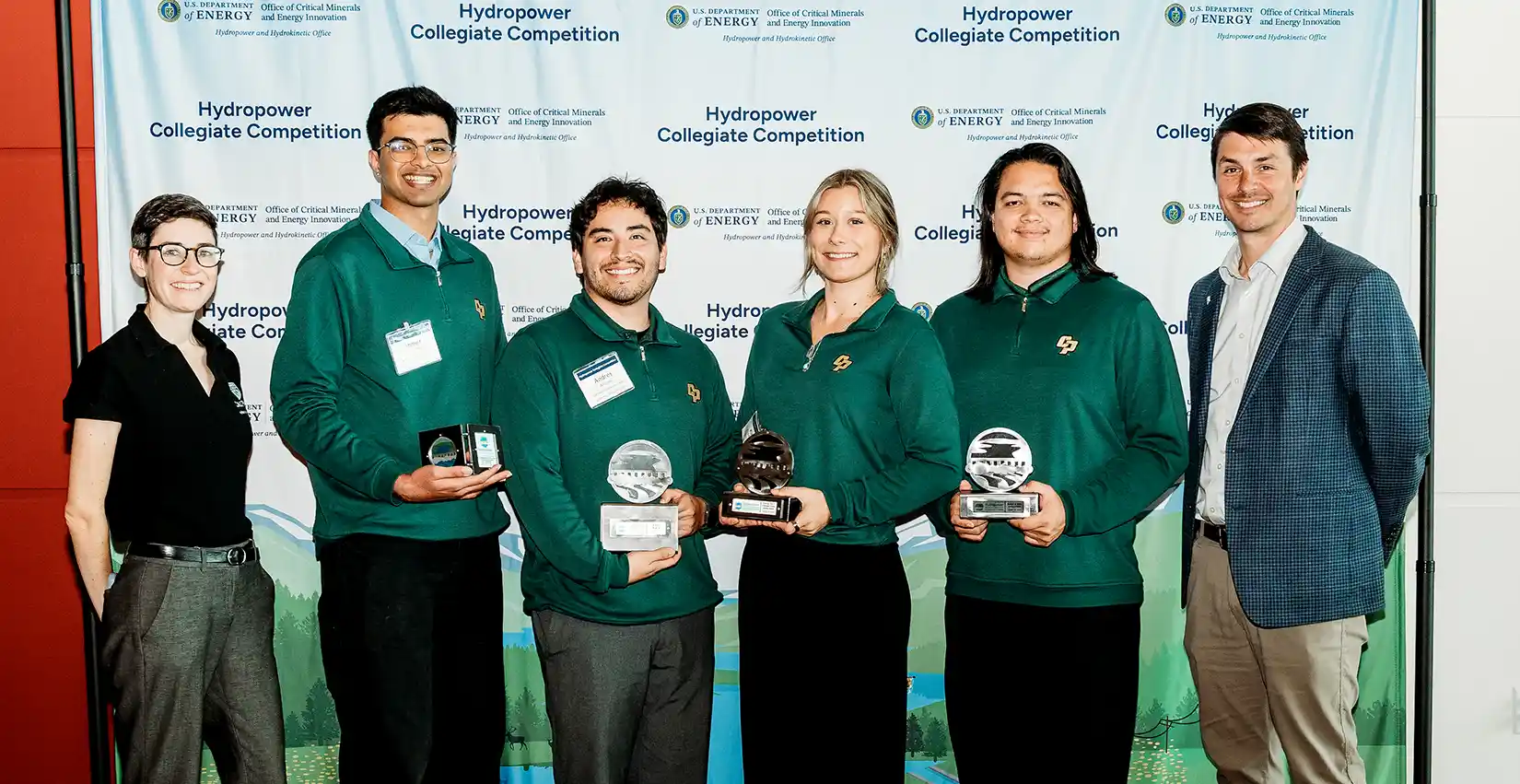 calpoly A photo of six people posing in front of a Hydropower Collegiate Competition sign. Four of the six are wearing green team jackets and holding awards in their hands.