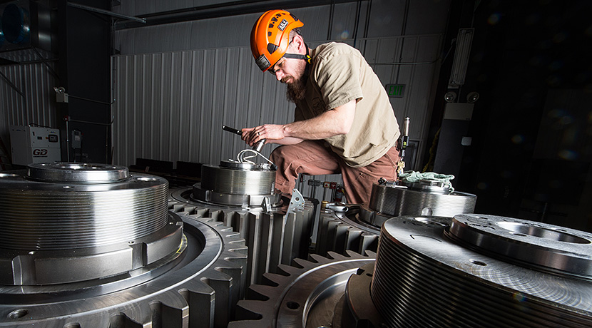 a man wearing an orange helmet and measuring a pin on one of three large gears, which are part of a drive train.