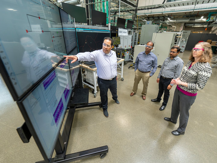 Four people stand around a series of flat-screen monitors as one points at an object on one of them.