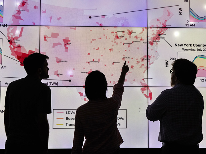 Silhouettes of three people pointing at a flat-screen monitor with a map of the United States.
