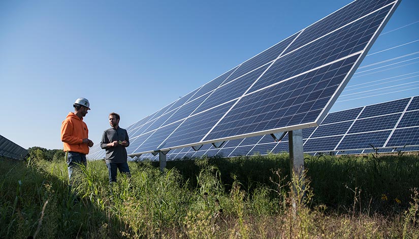 Two people standing next to large, ground-mounted photovoltaic arrays.