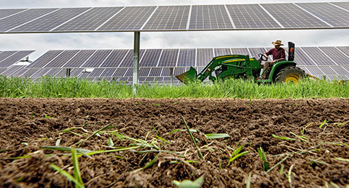A farmer drives a tractor beneath a solar array with tilled dirt in the foreground.