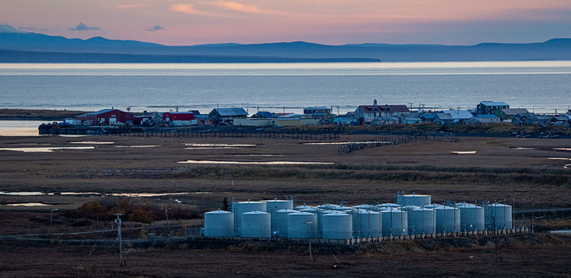 Buildings and homes line a waterfront with tanks in the foreground and mountains in the background