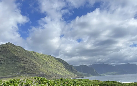 A man looks out at green mountains on the coastline under blue sky with clouds.
