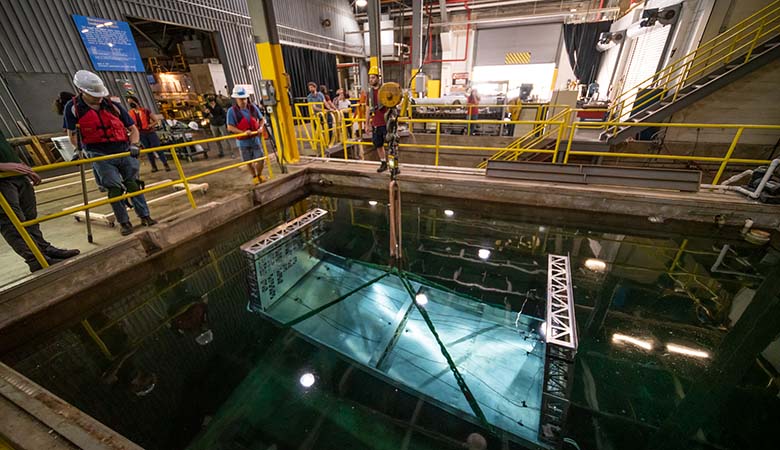 People in hard hats stand behind protective railing, looking down into a pit where a device is lowered by crane into water.